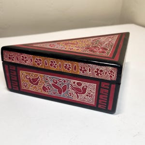 May include: A triangular black box with a hinged lid. The box is decorated with red, white, and yellow floral and bird designs. The box is likely made of wood and has a glossy finish.