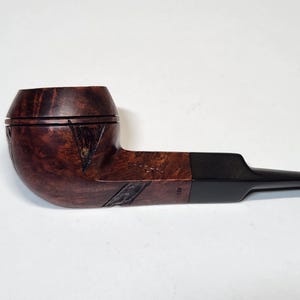 May include: A handcrafted tobacco pipe with a dark brown, curved bowl and a black stem. The bowl has a carved design and a rich, wood-grain texture. The pipe is designed for smoking and is a classic accessory.