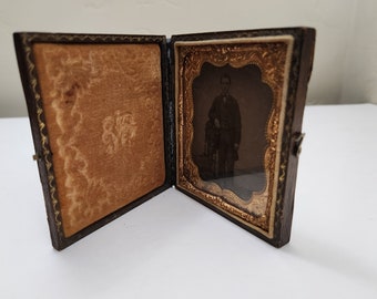 May include: A vintage, wooden, hinged photo frame with a velvet lining and a gold-trimmed, ornate frame surrounding a black and white photograph of a man in a suit.
