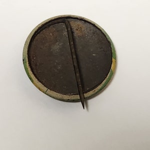 May include: The back of a round button with a metal pin. The button has a green and yellow border and a brown center.