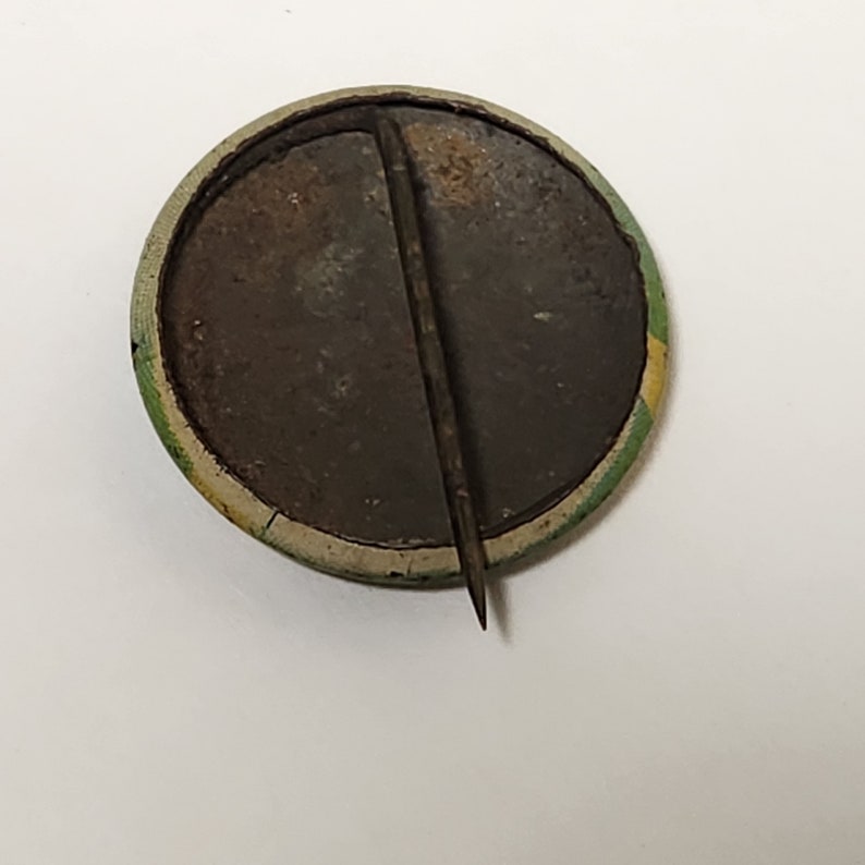 May include: The back of a round button with a metal pin. The button has a green and yellow border and a brown center.