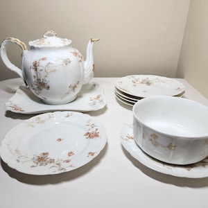 May include: A white porcelain tea set with floral accents. The set includes a teapot, plates, a bowl, and saucers. The teapot and plates have a scalloped edge. The floral design features brown and tan colors.