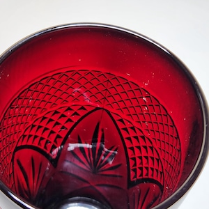 May include: Close-up of a ruby red glass with a detailed, geometric pattern etched into the interior. The glass has a dark rim and a decorative design at the base, showcasing a vintage aesthetic.