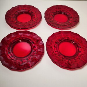 May include: Four ruby red glass plates with a ruffled edge and a central circular design. The plates have a detailed embossed pattern around the rim. The plates are arranged on a white surface.