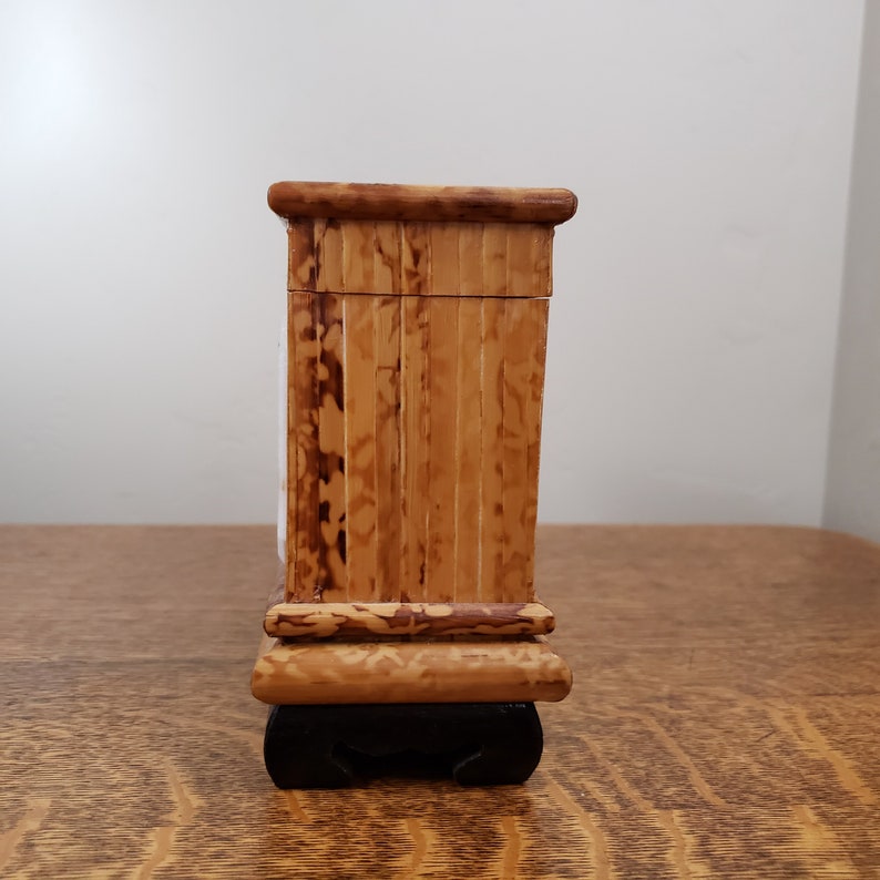 May include: A small wooden box with a brown and tan finish. The box has a hinged lid and sits on a black base.