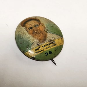 May include: A vintage green, yellow, and white button featuring a portrait of a man with the text "GEO. EARNSHAW PHILA. "ATHLETICS" 38".