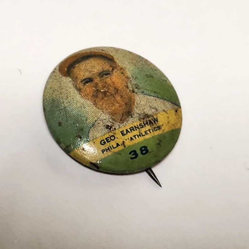 May include: A vintage green, yellow, and white button featuring a portrait of a man with the text "GEO. EARNSHAW PHILA. "ATHLETICS" 38".