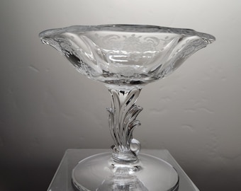 May include: A clear glass candy dish with a scalloped edge and an etched floral design. The dish sits on a pedestal with a decorative scroll design.