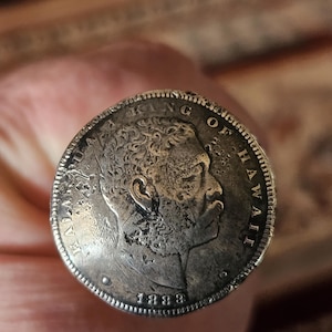 May include: A close-up of a silver coin featuring a profile of a man with the text "KING OF HAWAII" and the year "1883". The coin has a detailed design and is held in a person's hand.