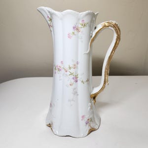 May include: A white porcelain pitcher featuring a fluted body with a scalloped rim and base. The exterior is decorated with small, delicate pink and purple floral sprays. A curved handle with ornate gold-painted detailing accents the side.