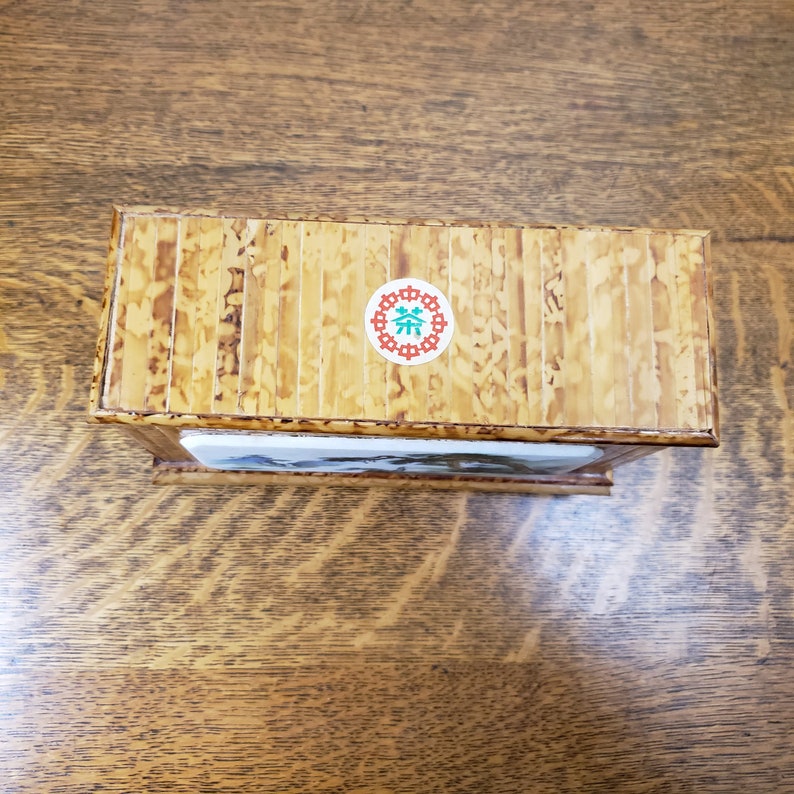 May include: A wooden box with a light brown finish and a sticker on the top that says "茶" in a green circle with a red border. The box has a decorative design on the front.