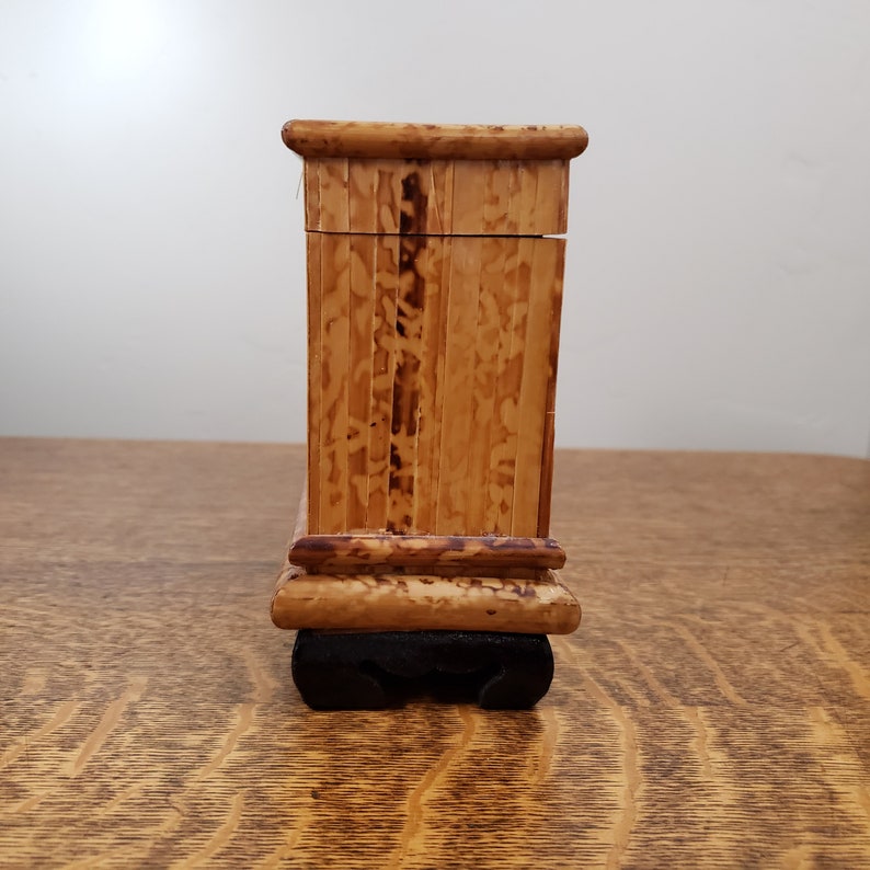 May include: A small, rectangular wooden box with a lid. The box is made of light brown wood with a dark brown base. The box is sitting on a wooden surface.
