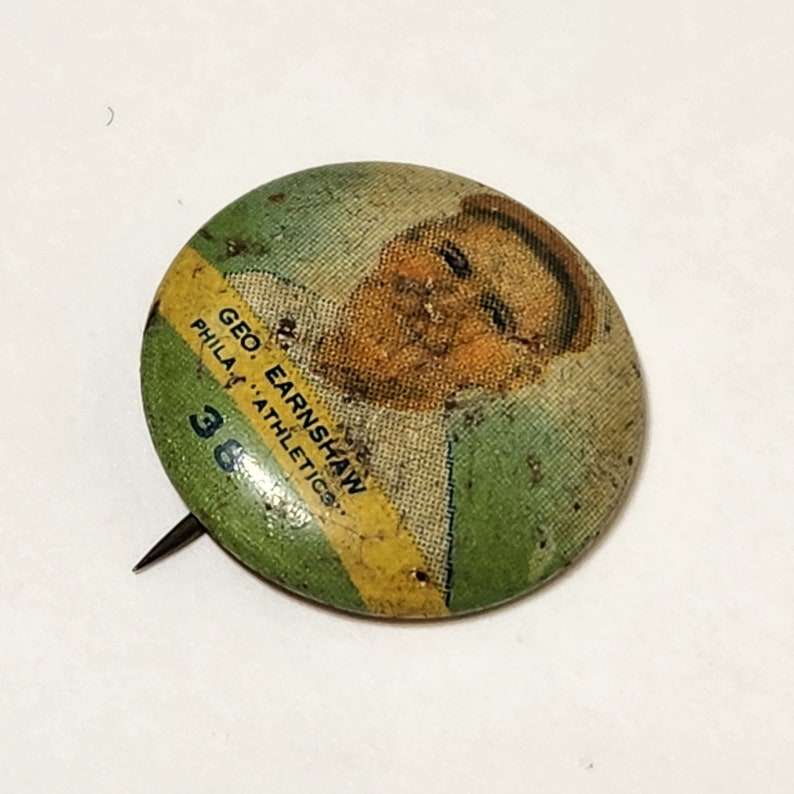 May include: A vintage green and yellow button featuring a photo of baseball player Geo. Earnshaw, Phila. Athletics, #38.