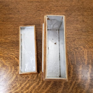 May include: Two wooden boxes with white interiors. The larger box is taller and has a rectangular shape. The smaller box is narrower and has a rectangular shape. Both boxes are open and empty.