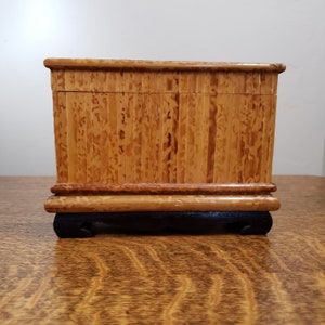 May include: A small wooden box with a natural wood finish. The box is made of thin strips of wood that are arranged vertically. The box has a black base with feet.