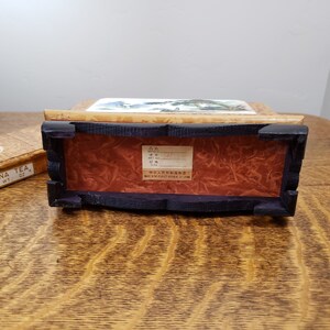 May include: A wooden box with a painted scene on the top. The box is closed and has a label on the bottom that reads "Made in the People's Republic of China".