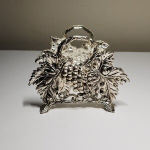 May include: Silver napkin holder with a grapevine design. The holder is shaped like a basket with a handle and has a detailed grape design.
