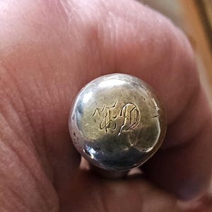 May include: A close-up of a silver, spherical object with an engraved design. The design appears to be initials or a monogram. The object is slightly tarnished, showing signs of age.