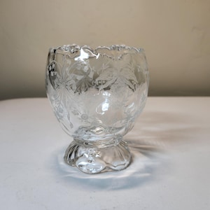 May include: Clear glass bowl with a floral etched design. The bowl has a rounded shape with a scalloped rim and sits on a pedestal base. The etched pattern features leaves and flowers. The bowl is empty and sits on a white surface.