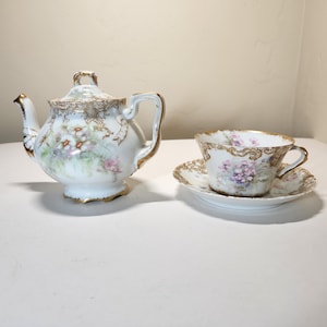 May include: A white porcelain tea set with gold trim, featuring a teapot, teacup, and saucer. The set is decorated with floral patterns in shades of pink, purple, and green. The teapot has a lid and a curved handle.