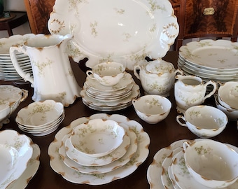 May include: A collection of antique white porcelain dishware with a delicate floral pattern in green and gold. The set includes plates, bowls, cups, a pitcher, and serving platters, arranged on a dark wooden surface.