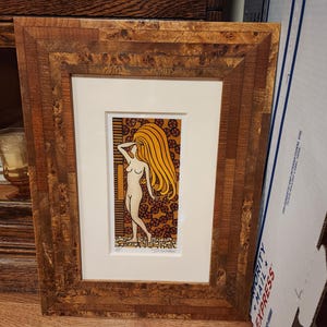 May include: Framed art print featuring a nude figure with long, flowing yellow hair. The artwork is set against a patterned background with a brown and gold color scheme. The frame is made of wood with a natural finish.