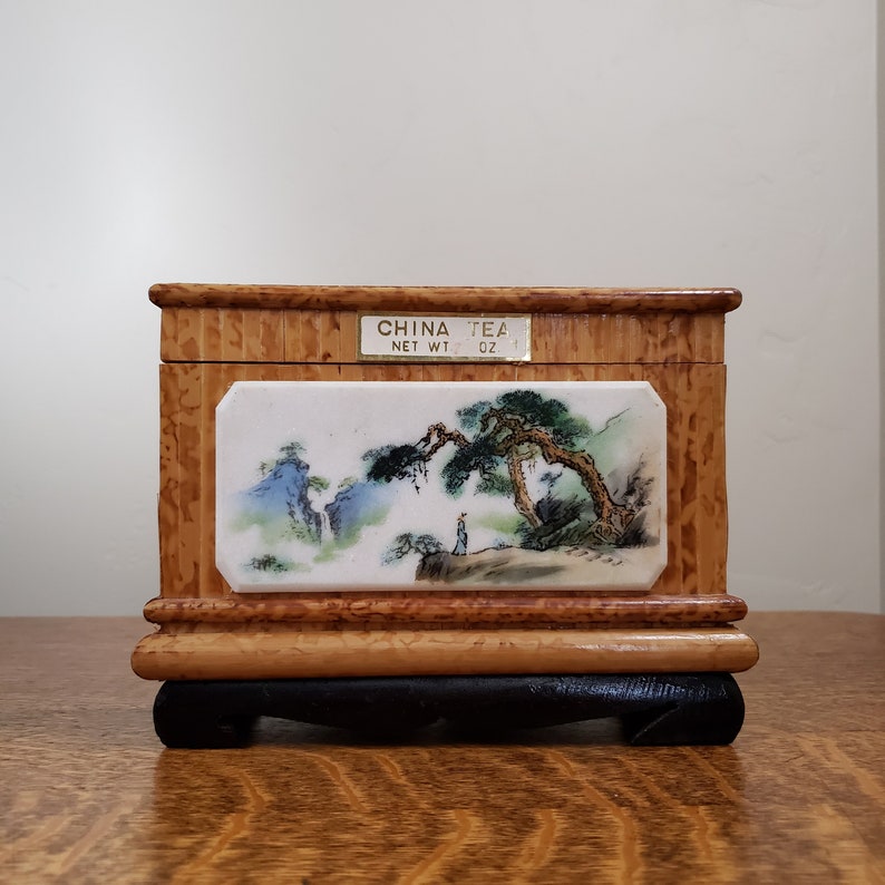 May include: A wooden tea box with a painted scene of a mountain landscape and a person standing on a path. The box has a label that reads "CHINA TEA NET WT. 1 OZ."