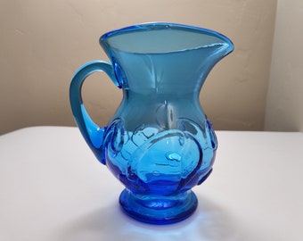 May include: A small, blue glass pitcher with a textured design. The pitcher has a handle and a round base.