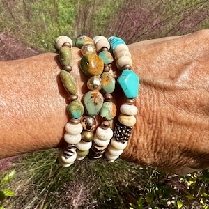 May include: A stack of five bracelets with turquoise, brown, gold, and white beads. The bracelets are layered on a person's wrist.