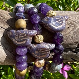 May include: Three bracelets with purple amethyst beads, gold beads, and purple bird charms. The bracelets are stacked on a wooden surface.