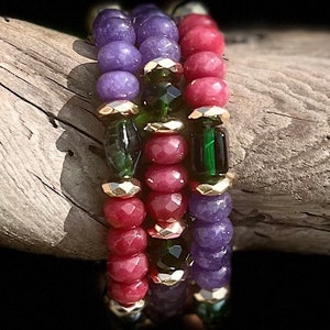 May include: Three stacked bracelets with purple, red, and green gemstone beads. The beads are separated by gold spacers.