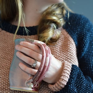 Puede incluir: Una persona sosteniendo una taza de cerámica con un esmalte azul claro, rosa y marrón. La taza tiene un asa texturizada única. La persona lleva un suéter rosa y azul.