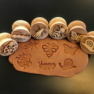 May include: Wooden insect-themed stamps with round handles. The stamps feature designs of a ladybug, caterpillar, insect, bee, butterfly, and snail. The stamps are pressed into a brown clay, creating impressions of the insects.
