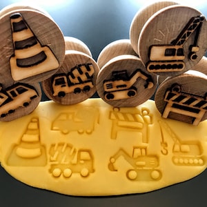 May include: Wooden construction-themed stamps with various vehicle designs, including a traffic cone, dump truck, cement mixer, excavator, crane, and road barrier. The stamps are pressed into yellow playdough, creating impressions of the same vehicles.