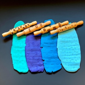 May include: Four wooden rolling pins with different designs. The rolling pins are on top of three different colored play doughs. The play doughs are imprinted with the designs from the rolling pins. The designs include letters, numbers, and shapes.