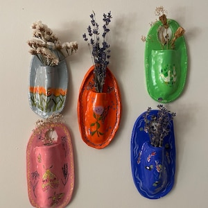 May include: Five oval-shaped ceramic wall vases in light blue, orange, green, pink, and dark blue. Each vase features hand-painted designs including flowers, butterflies, moths, geese, and koi fish, and contains dried floral arrangements.