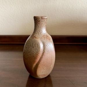 May include: A small ceramic bud vase with a narrow neck and a rounded, bulbous body. The surface features a textured, speckled finish in shades of tan, beige, and earthy brown, with a subtle curved indentation running vertically along the front side.
