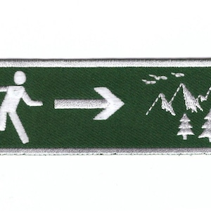 Op de afbeelding: Groene rechthoekige geborduurde patch met een wit rennende figuur die met een pijl naar rechts wijst. De achtergrond toont witte bergen en bomen.