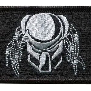 Könnte beinhalten: Ein schwarzer gestickter Aufnäher mit einer weißen Umrandung eines Predator-Alien-Kopfes.
