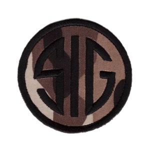 May include: A round, black embroidered patch with the letters "SIG" in a circle on a camouflage background.