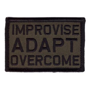 May include: Olive green rectangular patch with black embroidered text: IMPROVISE, ADAPT, OVERCOME. The patch has a black border and is likely designed to be sewn or attached to clothing or gear.