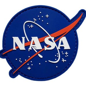 Könnte beinhalten: Ein blauer Aufnäher mit dem NASA-Logo. Das Logo zeigt das Wort "NASA" in Weiß, eine weiße Umlaufbahn und einen roten Schwung. Weiße Sterne und Pluszeichen sind um das Logo herum verstreut.