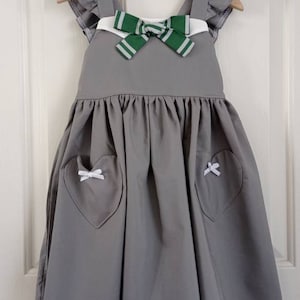 May include: A gray sleeveless dress with a white collar and a green and white striped bow. The dress has two heart-shaped pockets with white bows on each side.