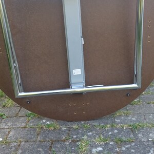 Może przedstawiać: Sp&oacute;d okrągłego, brązowego stołu z chromowaną, składaną ramą. St&oacute;ł ma srebrny metalowy słupek wsporczy i etykietę na słupku.