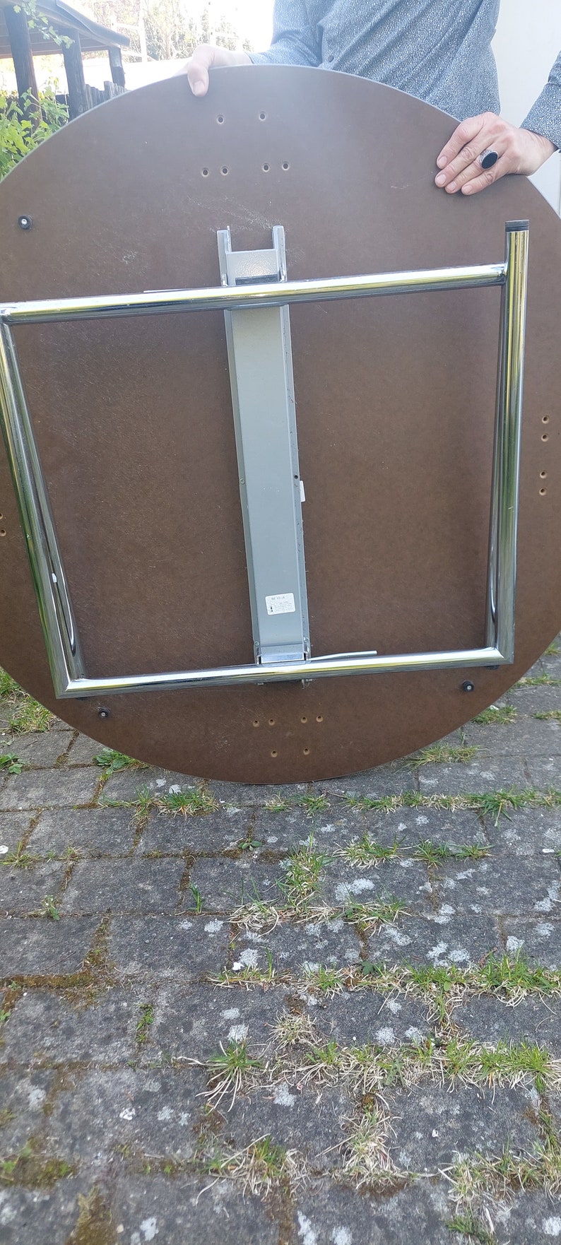 Może przedstawiać: Sp&oacute;d okrągłego, brązowego stołu z chromowaną, składaną ramą. St&oacute;ł ma srebrny metalowy słupek wsporczy i etykietę na słupku.