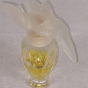 Puede incluir: Un frasco de perfume de vidrio transparente con un tapón blanco en forma de flor. El frasco tiene un diseño texturizado y contiene un líquido amarillo.