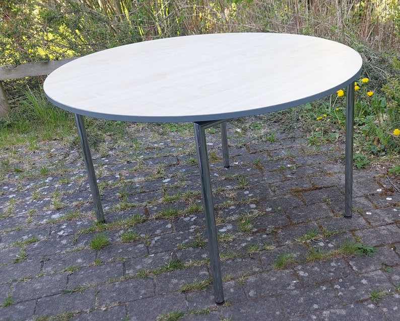 Może przedstawiać: Okrągły, biały st&oacute;ł z srebrną metalową ramą. St&oacute;ł stoi na brukowanym patio.