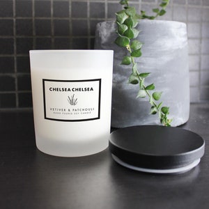 May include: A white frosted glass candle jar with a black label that reads "Chelsea Chelsea Vetiver & Patchouli Hand Poured Soy Candle". The candle is sitting on a black surface next to a black lid.