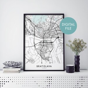 May include: Black and white line art map print of Bratislava, Slovakia in a black frame. The print is on a white background and has the city name and country name at the bottom. The print is on a white shelf with a black vase and a black geometric bowl.