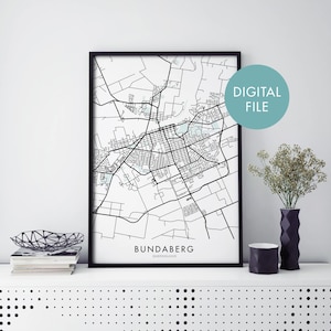 May include: Black and white line art map print of Bundaberg, Queensland, Australia. The map is framed in a black frame and is displayed on a white shelf with a black vase and a black and white bowl.